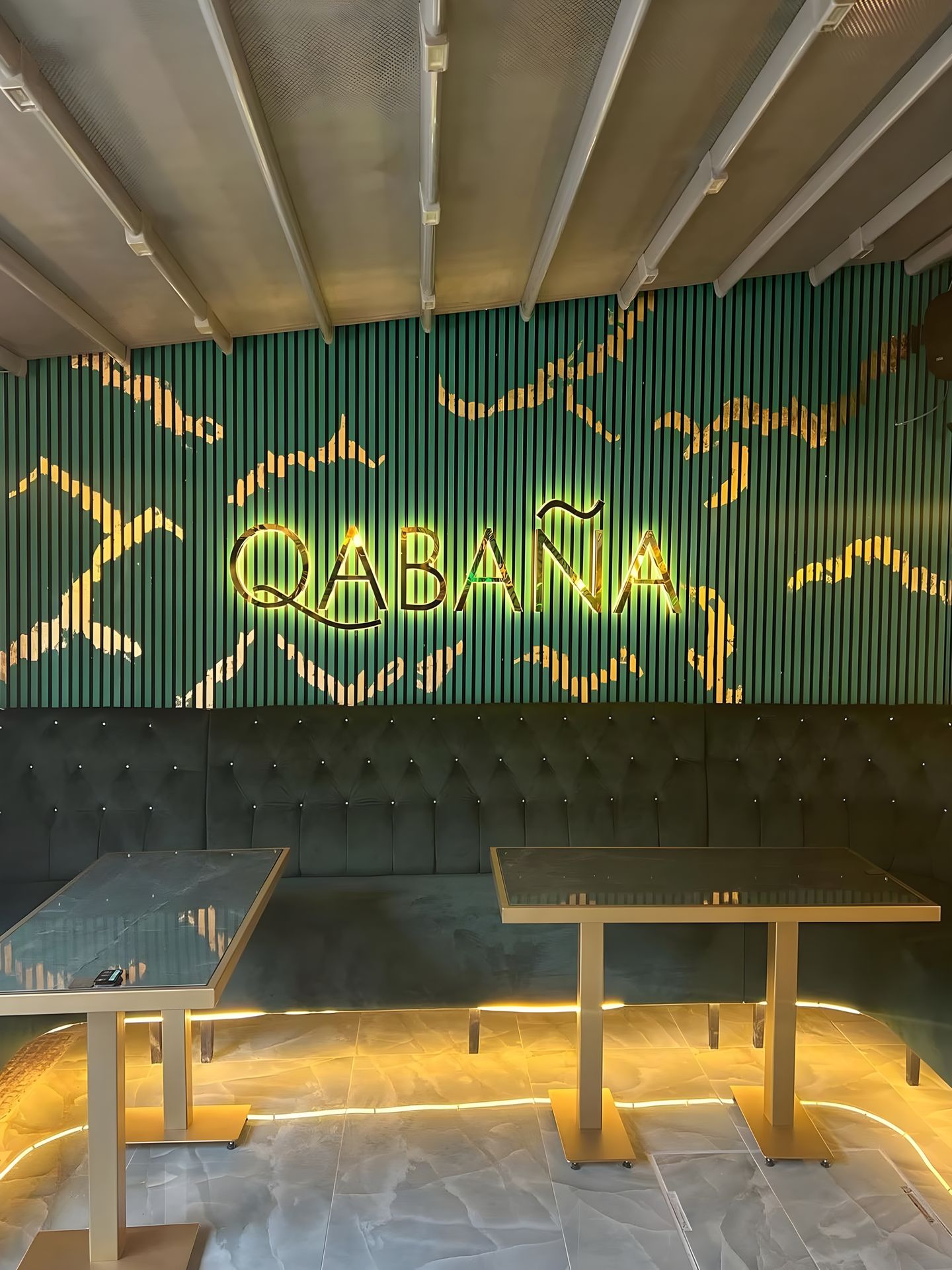 Qabaña: the illuminated QABAÑA wordmark on deep green fluted panelling with gold veining, a buttoned green banquette and brass-framed glass tables lit from beneath under a pale canopy.